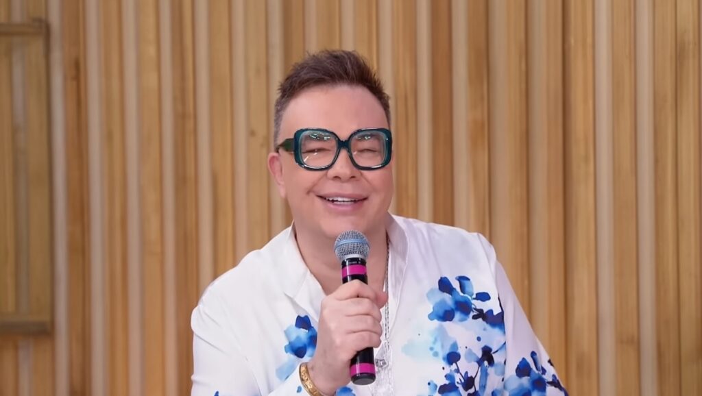 Jornalista Felipeh Campos sorrindo em estúdio de TV com banner de notícia urgente sobre saúde.